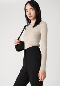 Top de punto acanalado beige con cuello alto y silueta ajustada, combinado con pantalones negros y un pequeño bolso negro.