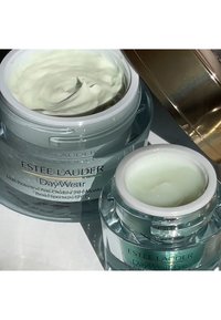 Zwei Glasbehälter mit Estée Lauder Creme. Der größere Behälter ist hellgrün mit einem weißen Deckel, der mit "DayWear" beschriftet ist. Der kleinere Behälter ist ähnlich und trägt die Aufschrift "DayWear Eye".
