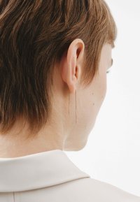 Pendientes colgantes en tono dorado con cadenas delgadas y rectas. Diseño ligero, que muestra una estética minimalista. Llevados por una persona con cabello corto.