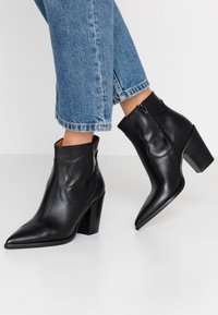 Botas de tobillo de cuero negro con punteras afiladas y tacones cuadrados, combinadas con jeans de mezclilla azul sobre un fondo blanco.