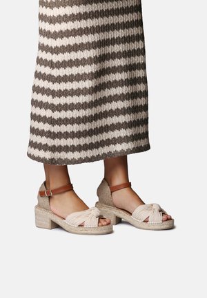 Austi smėlio spalvos basutės su blokiniu kulnu, su mazgu priekyje ir ruda čiurnos dirželiais, dėvimos su rudu ir baltu dryžuotu megztu sijonu.