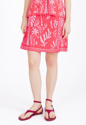 Kobieta w czerwonej spódnicy z różowymi tropikalnymi wzorami i czerwonych sandałach na kostkę, stojąca na białym tle.