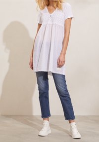 Femme portant une tunique en dentelle blanche sur un jean bleu et des baskets blanches montantes, se tenant sur un sol clair devant un mur uni.