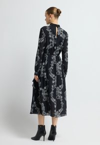 Schwarzes Langarmkleid mit weißem Paisley-Muster, tailliert, mit Schlüssellochausschnitt am Rücken und fließendem Rock, kombiniert mit schwarzen Stiefeletten mit hohen Absätzen.