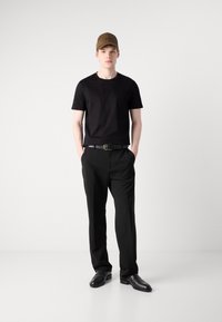 Black t-shirt, black trousers, and black shoes. Brown cap with logo. Model stands with hands in pockets against a plain white background.