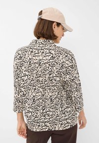 Blusa a maniche lunghe in tessuto beige con motivo animalier leopardato nero, dotata di colletto e dettagli con bottoni nella parte posteriore del collo.