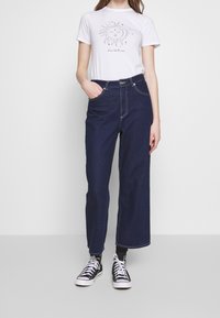 Camiseta blanca con un gráfico del sol, combinada con unos pantalones vaqueros de pierna ancha de tiro alto en azul oscuro y zapatillas en blanco y negro.