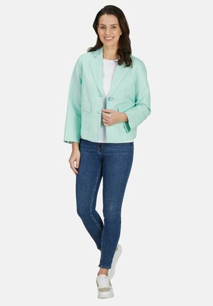 Frau lächelt, trägt einen hellgrünen Blazer, weißes Hemd, blaue Jeans und weiße Sneakers, steht vor einem einfachen hellen Hintergrund.
