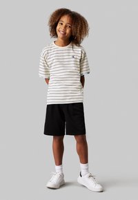 Camicia a maniche corte a righe color crema con linee blu e navy; abbinata a pantaloni corti neri e sneakers bianche. Vestibilità casual con design classico.