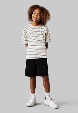 Camicia a maniche corte a righe color crema con linee blu e navy; abbinata a pantaloni corti neri e sneakers bianche. Vestibilità casual con design classico.
