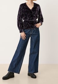 Blusa avvolgente in velluto viola scuro con motivi irregolari e maniche lunghe, abbinata a jeans in denim blu a gamba larga e stivaletti neri.