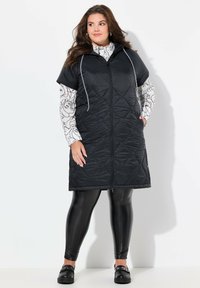 Černá prošívaná bezrukávová bunda s kapucí a předním zipem, nošená přes vzorovaný dlouhý rukáv, spárovaná s černými legínami a botami.