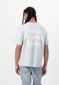 Light blue cotton t-shirt with short sleeves, featuring large white text on the back reading "Studio Originals Edition." Casual design.