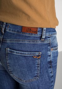 Pantalones vaqueros azules de mezclilla con una etiqueta de cuero marcada con "LTB". Presentan un bolsillo trasero con costuras claras y una textura desgastada.