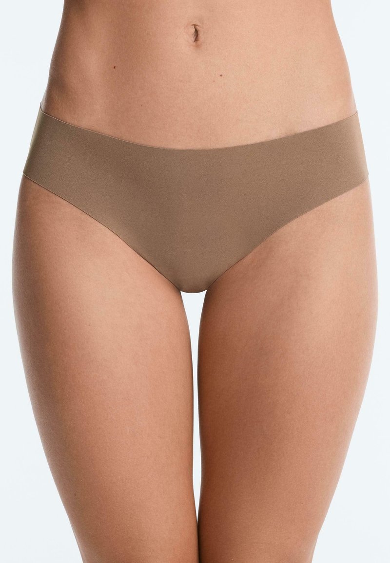 Femme portant un sous-vêtement hipster sans couture couleur chair, présenté de la taille jusqu'aux cuisses sur un fond blanc uni.