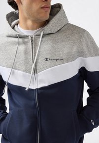 Uomo che indossa un hoodie con zip caratterizzato da blocchi di colore grigio, bianco e navy, logo Champion sul petto e sulla manica, con le mani nelle tasche, che guarda a destra.