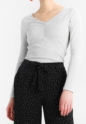 Frau trägt ein eng anliegendes, hellgraues, geripptes Langarmshirt und hoch taillierte schwarze Hose mit kleinen weißen Mustern und einem gebundenen Gürtel.