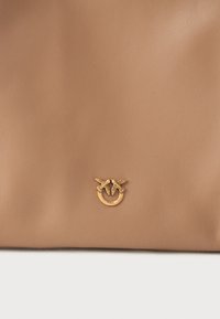 Tan spalvos odinė rankinė, kuri centre turi auksinės spalvos emblema su gėlių akcentais. Lygus paviršius, minimalus siuvinėjimas ir paprastas dizainas.