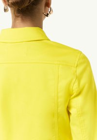 Yellow denim jacket with a collar and back yoke detail. Features visible stitching and a smooth texture. Gold geometric earrings are partially visible.