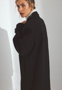 Čierny vlnený kabát s vysokým golierom, nadrozmerná silueta a detaily na gombíky. Hladká textúra s bočnými vreckami, zobrazený v prirodzenom svetle.