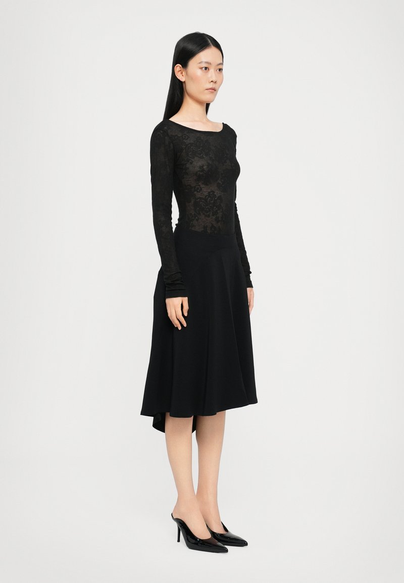 Abito nero con un corsetto in pizzo aderente e gonna svasata. Maniche lunghe, scollatura alta e orlo posteriore leggermente più corto. Indossato con tacchi neri.