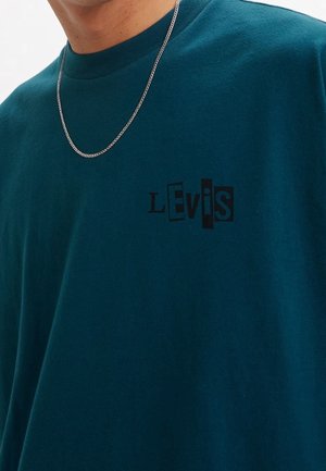 Persona con camisa verde azulado oscuro con logo negro estilizado "LEVI'S" en el pecho y un collar de cadena plateada fina.