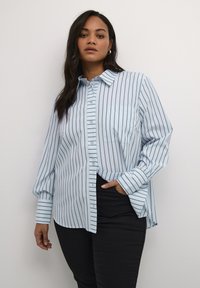 Camisa azul clara de botones con rayas oscuras verticales, cuello, bolsillo frontal y mangas largas; diseñada con un corte relajado y detalles de puño.