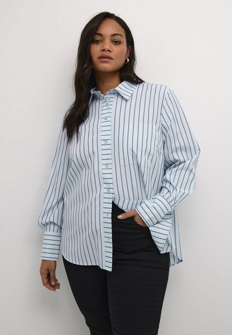 Camisa azul clara de botones con rayas oscuras verticales, cuello, bolsillo frontal y mangas largas; diseñada con un corte relajado y detalles de puño.