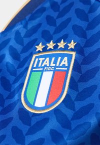 Blue football jersey fabric features a textured leaf pattern. The embroidered badge displays the Italian flag with four gold stars above.