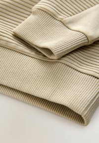 Vribrovaná béžová pletenina svetru s texturovaným, vícevrstvým designem. Manžety a lem mají podobný žebrovaný vzor. Měkký, útulný materiál.