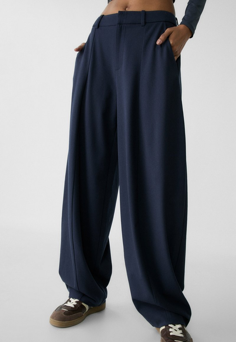 Personne portant un pantalon large ample bleu marine avec poches latérales et des baskets marron avec des lacets blancs.