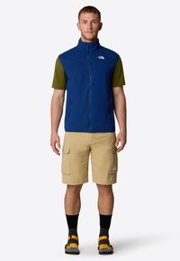 Gilet di pile blu con colletto alto, camicia verde a maniche corte, pantaloni cargo beige, calze nere e sandali arancioni su una persona.