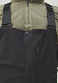 Sorte vandtætte bib overalls med justerbare sorte stropper, brystlomme med lynlås og fremtrædende knapdetaljer over en struktureret olivengrøn fleece-top.
