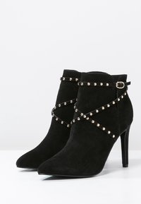 Botines de ante negros con puntera afilada, adornados con tachuelas doradas y correas entrelazadas, que cuentan con un tacón alto y delgado.