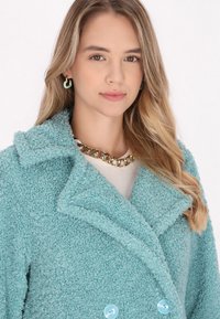 Cappotto vaporoso color teal con colletto a scialle, design doppiopetto e due bottoni azzurri. Indossato sopra un top chiaro, accessorato con una catena dorata.