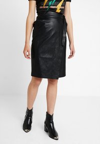 Jupe enveloppante en cuir noir avec une texture lisse, dotée d'une fermeture à nouer sur le côté et d'une coupe droite, associée à des bottines noires.
