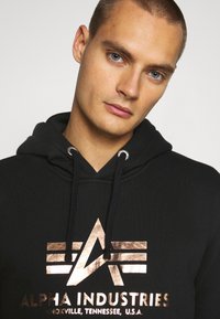 Felpa nera con cappuccio, caratterizzata da un logo e strisce in oro rosa metallico. Venti con hardware argentato, tessuto morbido, design semplice.