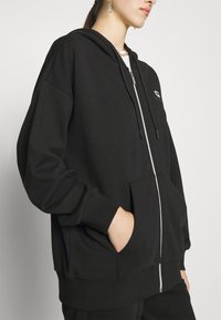 Musta vetoketjullinen huppari pehmeästä kankaasta, jossa on etutasku, säädettävät nyöritykset ja resorit hihansuissa. Rintakehällä logotyyppinen yksityiskohta.