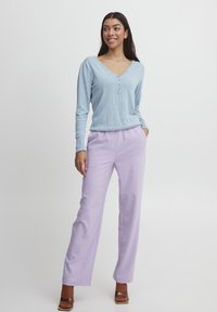 Haut bleu clair à manches longues avec un col en V et trois boutons ; associé à un pantalon large lavande et des chaussures à talons marron.