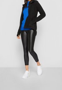 Svart fleecejacka med huva, blå stickad tröja, svarta glänsande leggings och vita sneakers, stående mot en enfärgad bakgrund.