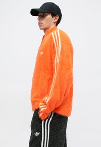 Oranžová chlpáčová bundu na zips so zelenobielym trojprúžkovým dizajnom, kombinovaná s čiernymi štruktúrovanými nohavicami s bielymi pruhmi a logom Adidas.