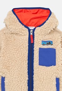 Bež fleecová bunda s modrou kapucňou a vreckom, which má červený zips a živou oranžovú podšívku. Na hrudi je zobrazené logo Patagonia.