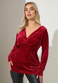 Czerwony aksamitny top z długim rękawem, z głębokim dekoltem w kształcie V i skręconym przodem. Gładka faktura, dopasowany krój i długie rękawy.