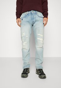 Jeans en denim bleu clair avec des accents déchirés, présentant des patches déchirés et une coupe droite, associés à des baskets noires et vertes.