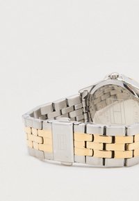 Ocelový hodinky se stříbrným a zlatým nerezovým řemínkem, kulatým pouzdrem a jednoduchým, přehledným ciferníkem. Obsahují sponu na zavírání.