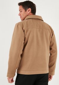 Beżowa kurtka z mieszanki wełny z klasycznym kołnierzem, dopasowanym krojem i kieszeniami wsuwanymi; posiada usztywniane ramiona i gładką teksturę.