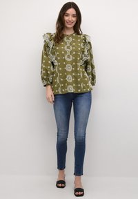 Olivgrön blus med vita blommönster, volangärmar och avslappnad passform, kombinerad med tighta blå jeans och svarta slidessandaler.