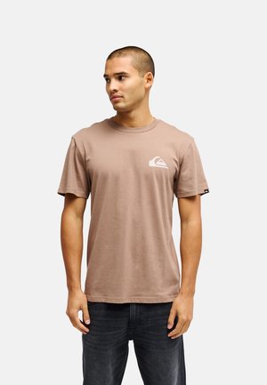 Jonge man met kort haar die een bruin T-shirt draagt met een wit logo op de borst en donkere jeans, kijkt opzij.