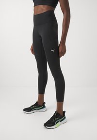 Leggins de sport noirs avec une taille haute, arborant un logo Puma discret. Portés avec des baskets noires rehaussées de détails verts.