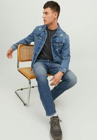 Chaqueta de mezclilla con detalles desgastados, combinada con una camiseta gris y jeans azules. Sentado en una silla de marco metálico con asiento de mimbre.